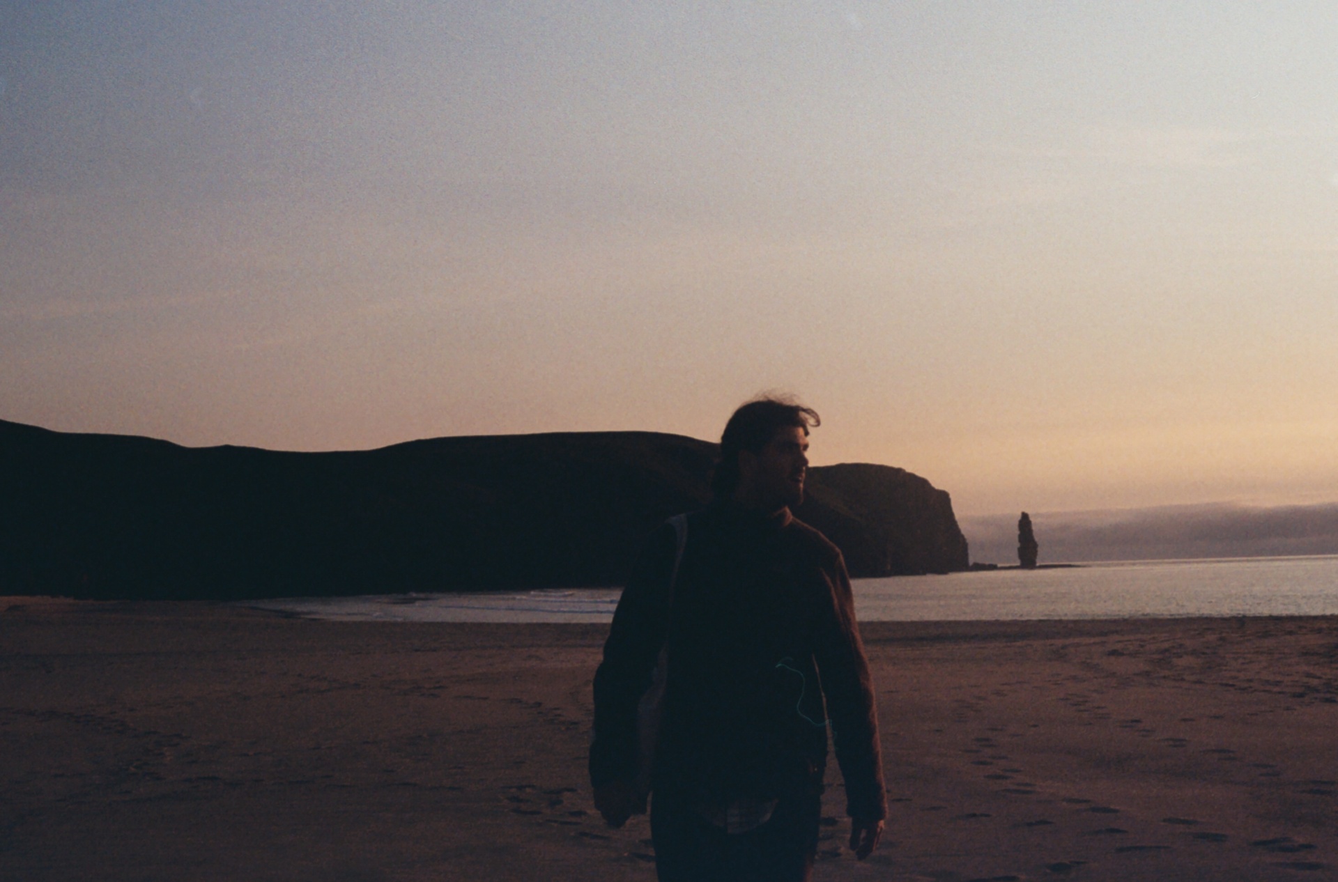 Joe walking on a beach at dusk, sea stack on the horizon
